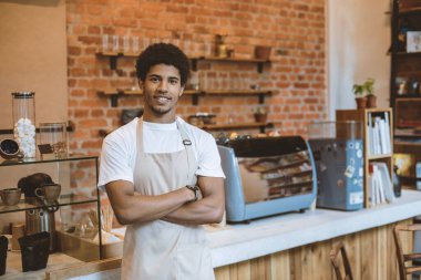 Kapatma sonrası modern kafenin açılışı. Önlüklü Afro-Amerikan genç adam kollarını kavuşturup modern tavan arası kafeteryasında kameraya bakıyor.