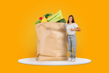 Genç bir kadın taze sebze ve meyvelerle dolu büyük kahverengi bir kesekağıdının yanında duruyor. Başarılı bir alışveriş deneyimini kutlarken, alışverişini yaparken gülümsüyor..