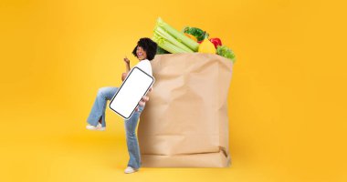 Bir müşteri, bir elinde taze ürünlerle dolu büyük bir kese kâğıdı taşırken elinde cep telefonu tutarak market alışverişiyle mutlu mesut ilgilenir..