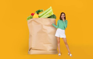 Taze sebze ve meyve dolu büyük bir kesekağıdının yanında duran bir müşteri elini kaldırır. Parlak arka plan, market alışverişine geçişi norm olarak vurguluyor.