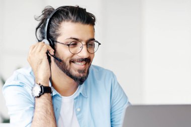 Gülümseyen Hintli genç adam dizüstü bilgisayarında kulaklık ve gözlük takıyor, online eğitimin ve uzaktan öğrenmenin tadını çıkarıyor, evde oturup bilgisayar ekranına bakıyor, boş alan arıyor,
