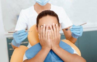 Çocuklarla çalışmak ve diş malzemelerinden korkmak. Korkmuş küçük melez çocuk, elleri diş sandalyesinde, yüzlerini kapatıyor, bin yıllık kadın doktorunun arkasında, koruyucu maske ve eldivenler takıyor.