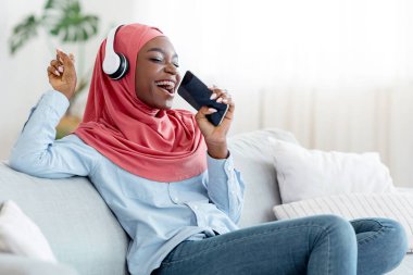 Mobil Karaoke. Neşeli siyahi Müslüman kadın, evde kablosuz kulaklıkla müzik dinleyerek, oturma odasındaki rahat koltukta dinlenerek, uzayı kopyalayarak, ellerinde akıllı telefonuyla şarkı söylüyor.