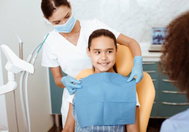 Modern diş kliniğinde hizmet. Beyaz önlüklü, koruyucu maskeli ve eldivenli hemşireye bakan, İspanyol tebessümlü, sandalyede gülümseyen küçük bir kıza peçete takan genç Afrikalı Amerikalı doktor.