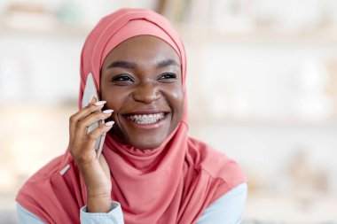 Telefon var. Neşeli siyahi Müslüman kız evde cep telefonuyla konuşuyor, keyifli sohbetler yapıyor, mobil iletişimin tadını çıkarıyor, diş telleriyle gülümsüyor, pembe başörtüsü takıyor, bedava uzay boşluğu açıyor.