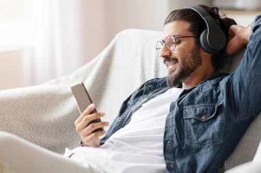 Rahat Hintli adam akıllı telefonuyla müzik dinliyor, kablosuz kulaklıkla evde dinleniyor, cep telefonundaki favori şarkıların tadını çıkarıyor, şarkı listesini düşürüyor, oturma odasındaki rahat koltukta dinleniyor.