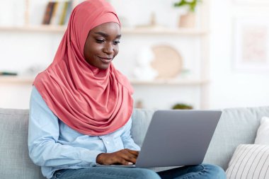 Çevrimiçi İletişim. Siyah Müslüman kadın başörtüsü içinde evde dizüstü bilgisayar kullanıyor, Afrikalı İslamcı kadın arkadaşlarıyla mesajlaşıyor, oturma odasında koltukta dinlenirken klavyede daktilo yazıyor, boşluğu kopyalıyor.