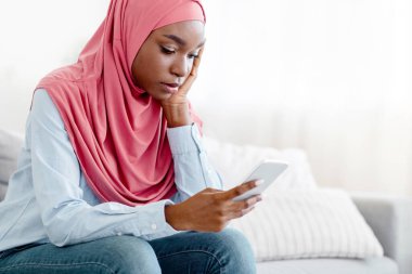 Üzgün, siyah, islami bir kadın evdeki akıllı telefondan mesaj okuyor oturma odasında kanepede oturuyor, depresif, tesettürlü, üzgün ve yalnız hisseden Afrikalı Müslüman bir kadın cep telefonu ekranına bakıyor, boşluğu kopyalıyor.