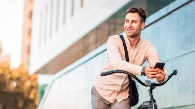 Neşeli adam dışarıda bisikletle poz veriyor. Modern ofis binasının yanındaki akıllı telefonlu genç hippi, kopyalama alanı