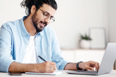 Genç Hintli iş adamı ofisteki dizüstü bilgisayarda çalışırken notlar alıyor, Batılı serbest çalışan adam internetten bilgi alıyor, planlama programı yapıyor, masada oturuyor, boşluğu kopyalıyor.