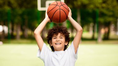 Kapalı arkaplanda basket topu atan kıvırcık saçlı çocuğun yakın plan portresi.