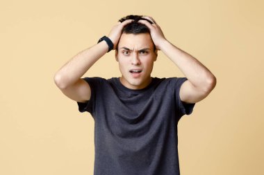 Hayal kırıklığı, başarısız olma, kaybetme ve çıldırma. Kahverengi tişörtlü, ağzı açık, başını eliyle tutan, bej arka planda izole edilmiş, stüdyo çekimi, serbest alan.