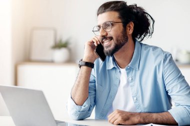 Genç Hintli erkek girişimci ofisinde cep telefonuyla konuşuyor, keyifli bir telefon görüşmesi yapıyor, iş görüşmesi için cep telefonunu kullanıyor, dizüstü bilgisayarla masada oturuyor, boşluğu kopyalıyor,
