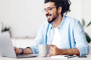 Serbest İşlerin Profesyonelleri. Olumlu Arap serbest çalışanı kahve içiyor ve merkez ofiste dizüstü bilgisayarda çalışıyor, Doğu Yakalı adam masada oturuyor ve klavyede daktilo yazıyor, uzaktan kumandayla eğleniyor, boş alandan keyif alıyor.