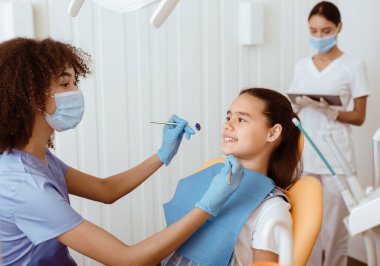 Afrika kökenli Amerikalı dişçi ve diş tedavisi. Mutlu genç doktor ve küçük kız iletişim kuruyor, Stomatolojistin portresi modern klinikte arka planda çalışan bir ekip, kopyalama alanı