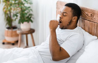 Uykulu Afro-Amerikan adam esneyerek uyanıyor. Sabahları yatakta oturuyor. Boşluğu Kopyala