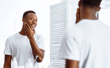 Evdeki modern banyoda, traş olurken aynanın yanında duran Afrikalı Amerikalı adam pürüzsüz bir yüze dokunuyor. Erkek Güzelliği ve Yüz Bakımı. Seçici odak