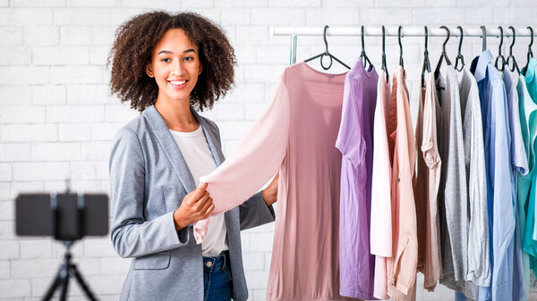 Модный блог, веб-бинар и онлайн-урок стиля. Улыбающаяся американка из Африки записывает видео для студентов на самоизоляцию во время карантина в интерьере гостиной рядом с стойкой с одеждой