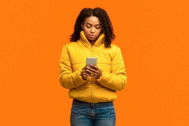 Ciddi Afrikalı Amerikalı Kız, Smartphone Kullanıyor, Kış Giysileri Giyiyor, Sarı Arkaplanda Duruyor, Stüdyo Çekimi. Çağrı Bekleniyor