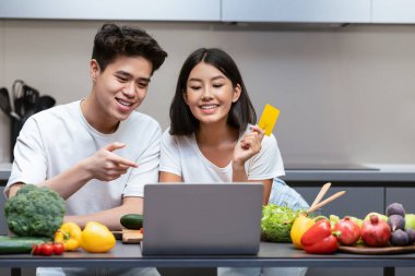 İnternetten alışveriş yapan Asyalı çift laptopta alışveriş yapıyor kredi kartı tutuyor modern mutfakta birlikte yemek pişiriyorlar. Aile Yemek Siparişi Hafta sonu İnternet Teslimat Servisi