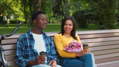 Genç bir Afro-Amerikalı çift parkta bir bankta oturup kahkahalarını içerken, bir buket çiçek tutuyor. Güneşli bir günde dışarıda eğleniyorlar..