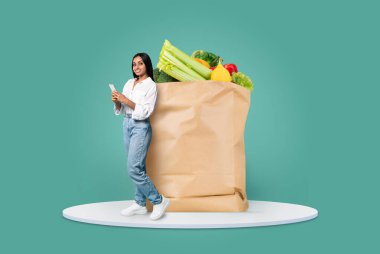 Bir müşteri taze sebze ve meyvelerle dolu büyük bir poşetin yanında duruyor. Telefonunu kontrol ederken gülümsüyor. Bu modern alışveriş alışkanlıklarını temsil ediyor..