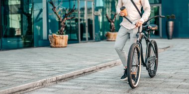 Genç bir adam bisikletine yaslanırken tesadüfen bir kahve fincanı tutuyor. Hafif giysiler içinde modern bir şehir caddesinde duruyor. Etrafı gösterişli binalar ve ekicilerle çevrili.