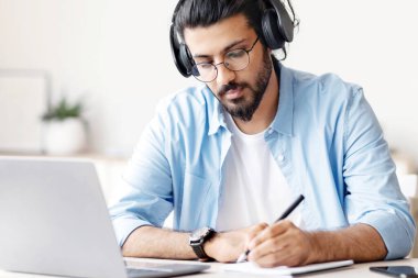 Mesafe öğrenme. Kulaklıklı genç bir Arap, evde dizüstü bilgisayarla ders çalışıyor ve not alıyor, Webinar 'ı izleyen yakışıklı bir doğulu ya da internetten eğitim alan ya da boş alan arayan bir adam.