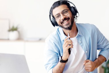 Telsiz kulaklıklı neşeli bir Arap adam, akıllı telefonuyla müzik dinliyor ve merkez büroda otururken şarkı söylüyor, neşeli bir Doğulu adam cep telefonunu mikrofon gibi kullanıyor ve kameraya gülümsüyor.