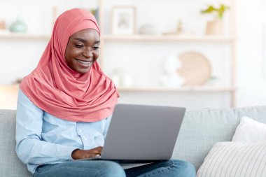 Uzaktan kumanda. Sırıtkan Afrikalı Müslüman kadın evde dizüstü bilgisayar kullanıyor, bilgisayarla koltukta oturuyor, serbest çalışmanın keyfini çıkarıyor, internette çalışırken bilgisayar ve ücretsiz alan ile koltukta oturuyor.