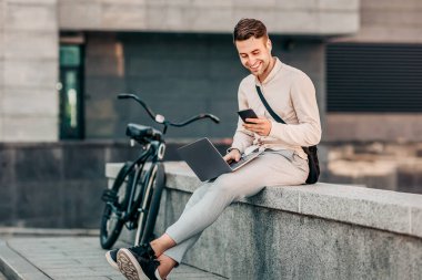 Bisikletli genç bir adam taştan bir çıkıntıda oturuyor, telefonunda kayarken dizüstü bilgisayarıyla çalışıyor.