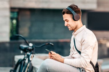 Genç bir adam sokakta dinleniyor, telefonunu kontrol ediyor ve kulaklık takıyor. Bisikleti yakınlarda park edilmiş. Güneşli bir gün ve müziğe dalmış, şehir yaşamının tadını çıkarıyor..