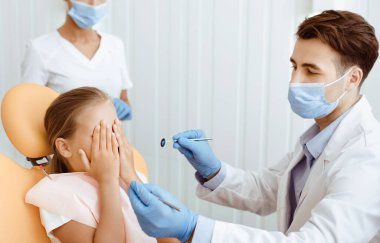 Modern diş hekimliğinde hastaların dişlerini muayene eden bir diş hekimi. Küçük kız korkmuş ve sandalyeye oturmuş modern stomatoloji kliniğindeki doktoru ziyaret ederken gözlerini kapatıyor. Maskeli dişçi tıbbi ekipmanları elinde tutuyor.