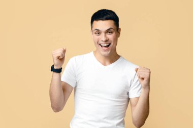 Heyecanlı mutlu öğrenci mutluluktan bağırıyor, en büyük zaferini ve zaferini kutluyor, yumruklarını sıkıyor, başarıya inanamıyor. Kazanan genç adam, bej arka planda izole edilmiş, serbest alan