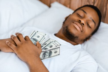 Siyah adam elinde para tutarak uyuyor. Para dolu banknotlara sarılıyor. Zenginlik ve evde yatıp zengin olma hayali kuruyor. Finansal Tasarruf ve Bütçe, Mali Güvenlik Konsepti. Seçici Odaklanma