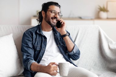 Yurt içi eğlence. Olumlu Arap adam arkadaşlarıyla telefonda konuşuyor ve evde kahve içiyor, keyifli bir telefon görüşmesi yaparken koltukta dinleniyor, rahat bir oturma odasında dinleniyor, mekanı kopyalıyor.