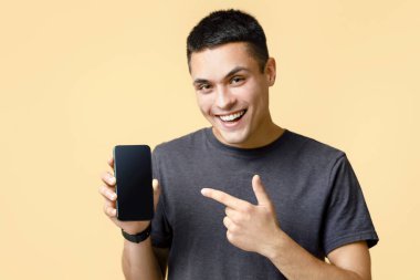 İnternette çalışmak, satış, modern cihazlar ve mobil uygulama reklamcılığı. Mutlu genç adam akıllı telefonu boş ekran ile işaret ediyor, açık kumlu arka planda izole edilmiş, stüdyo çekimi