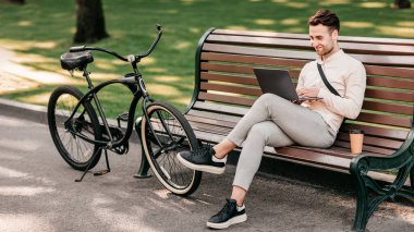 Genç bir adam parkta rahat bir şekilde oturuyor, dizüstü bilgisayarında yazıyor. Yanında bir bisiklet, bir kahve fincanı yerde oturup sakin bir hayat tarzını yakalar..