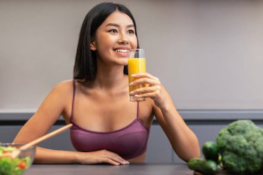 Detoks diyeti. Evdeki modern mutfaktaki masada oturan, sağlıklı sebze yemeği yapan, portakal suyu içen, heyecanlı Asyalı kadın. Beslenme ve Diyet, Zayıflama Konsepti