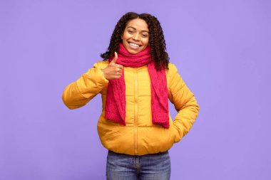 Siyah kadın kameraya gülümseyen, mavi stüdyonun arkasında duran, kışlık giysiler giyen bir şeyi onaylayan ve tavsiye eden hareketler yapıyor. 
