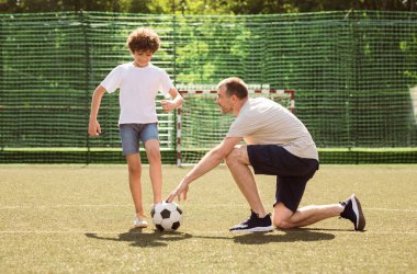 Açık havada egzersiz yapıyorum. Yetişkin bir adam küçük bir çocuğa futbol oynamayı öğretiyor, spor sahasında duruyor.