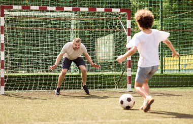 Aile maçı. Küçük çocuk futbol oynuyor, topa vuruyor, adam kalede duruyor.