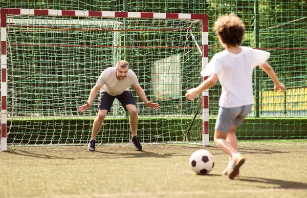 Aile maçı. Küçük çocuk futbol oynuyor, topa vuruyor, adam kalede duruyor.