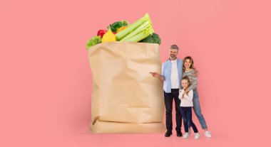 Bir aile, içi taze ürünlerle dolu büyük bir market poşetinin yanında duruyor. Neşeli ve modern yaşam tarzını yansıtan bir alışveriş deneyimiyle meşgul görünüyorlar..