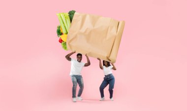 Bir çift enerjik bir şekilde taze sebze ve meyvelerle dolu büyük bir kesekağıdını parlak pembe arka planda tutuyor. Market alışverişinin mutluluğunu temsil ediyorlar..
