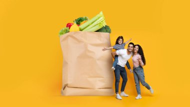 Üç kişilik bir aile, içi taze ürünlerle dolu büyük bir poşetle mutlu bir şekilde poz veriyor. Neşeli ifadeleri, canlı bir atmosferde birlikte alışveriş yapmanın olumlu bir deneyimini yansıtıyor..