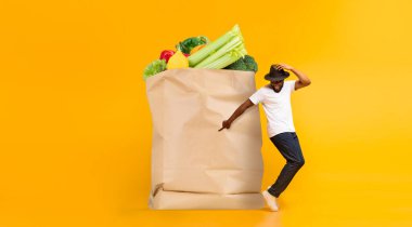 Bir müşteri renkli meyve ve sebzelerle dolu büyük bir kesekağıdının yanında duruyor, canlı bir pazar ortamında market alışverişinin ve teslimatın yeni normalliğini simgeliyor..