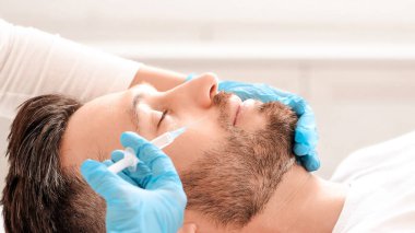 Yakışıklı sakallı adam estetik kliniğinde burun kıvırma dolgusu yaptırıyor. Plastik cerrah mavi eldivenler içinde zengin, orta yaşlı adama güzellik iğnesi yapıyor.