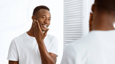 Evdeki modern banyoda dikilip aynaya baktıktan sonra tıraşlı suratını okşayan Afrikalı adam. Erkek Sabah Rutini, Yüz Derisi Kozmetik Konsepti. Panorama, Seçici Odaklanma