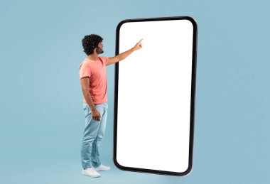 Havalı, kıvırcık Hintli genç adam, sıradan bir şekilde, mavi stüdyo arkaplanının üzerinde beyaz ekranda, büyük bir akıllı telefon ekranına dokunuyor, en yeni mobil uygulamayı kontrol ediyor, kopyalama alanı olan panorama, taklit alanı olan,